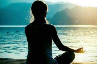 A woman facing the mountains and water at sunset in a Namaste yoga position